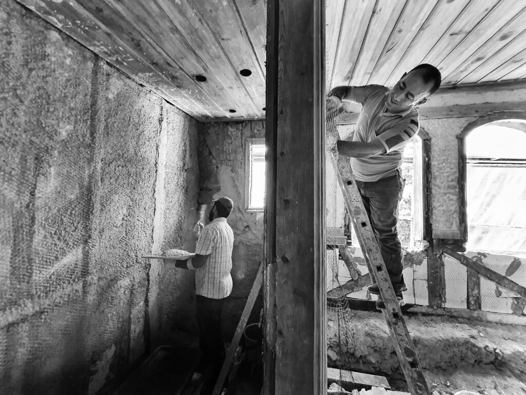 Two men renovating a room, one on a ladder applying plaster, showcasing teamwork and effort.