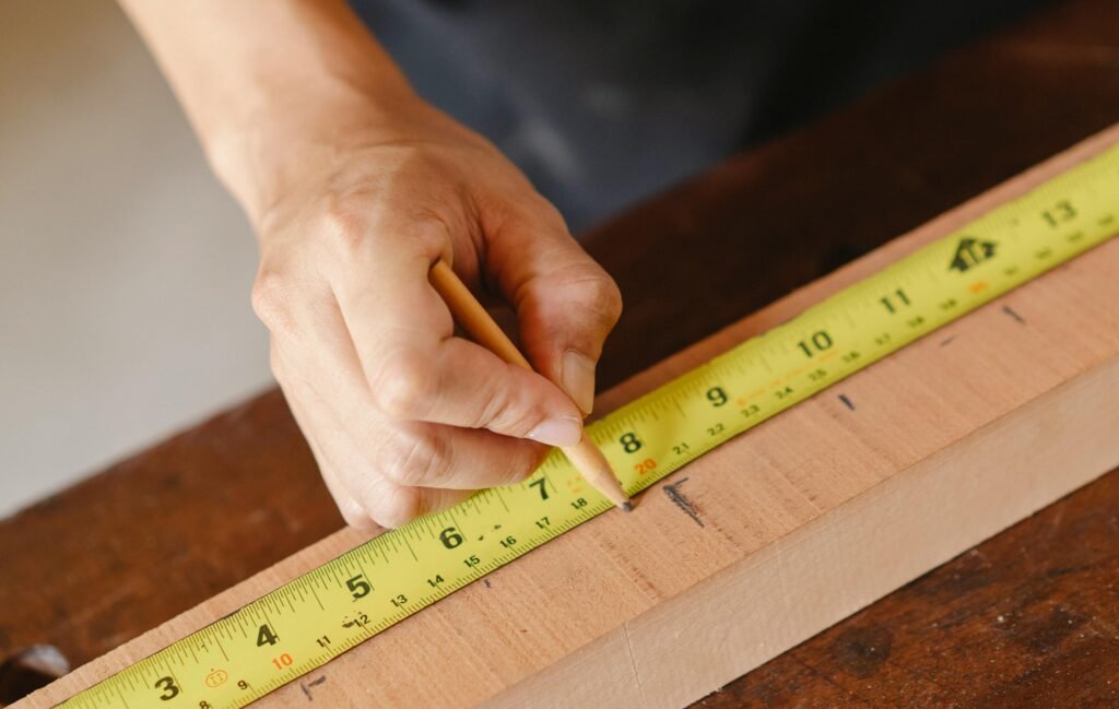 Detailed view of a hand measuring a wooden plank with a measuring tape and pencil in a carpentry setting.